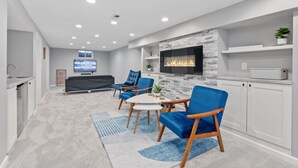 Un salon moderne avec une cheminée, des fauteuils bleus, un téléviseur et un coin cuisine.
