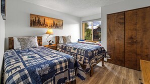 A bedroom with two beds, a wooden wardrobe, a painting of a forest, and a window with a view of a house.