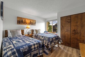 Ein Schlafzimmer mit zwei Betten, einem Holzschrank, einem Waldgemälde und einem Fenster mit Blick auf ein Haus.