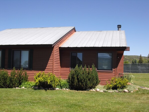 Une maison en bois rouge, surmontée d’un toit en métal, entourée d’une pelouse bien entretenue et de divers arbustes.