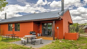 A red wooden cabin with a patio area featuring a grill, tables, and chairs.