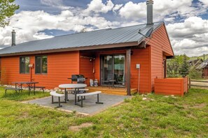 A red wooden cabin with a patio area featuring a grill, tables, and chairs.
