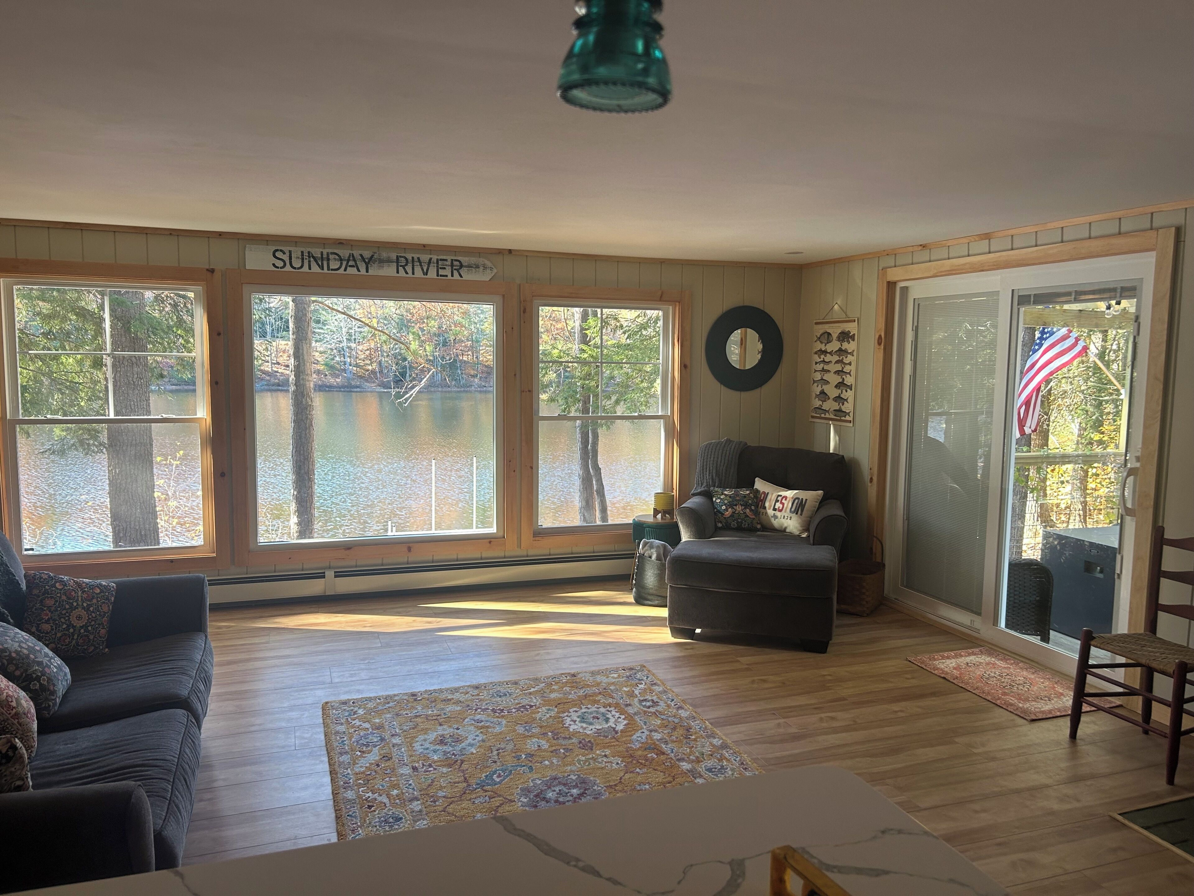A spacious living room with large windows overlooking a river, a comfortable seating area, and a view of the outdoors.
