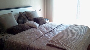 Un lit bien fait, avec une couverture matelassée et plusieurs oreillers, une table de chevet avec une lampe et une fenêtre avec des rideaux.