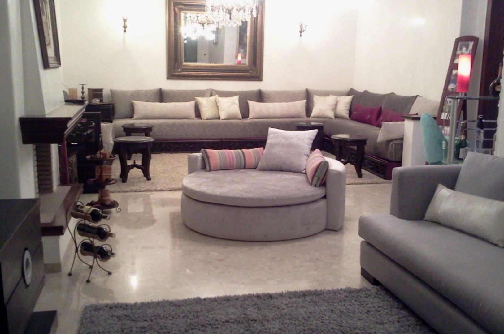 Un salon avec un canapé gris, un pouf rond et un fauteuil gris. On y trouve une table basse, une table d’appoint et une bibliothèque.