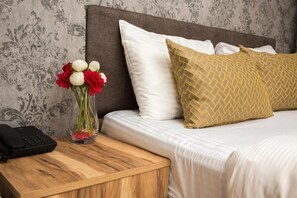 A neatly made bed with white and gold pillows, a bedside table with a vase of red and white flowers, and a black phone. - Honey Hotel (Istanbul)