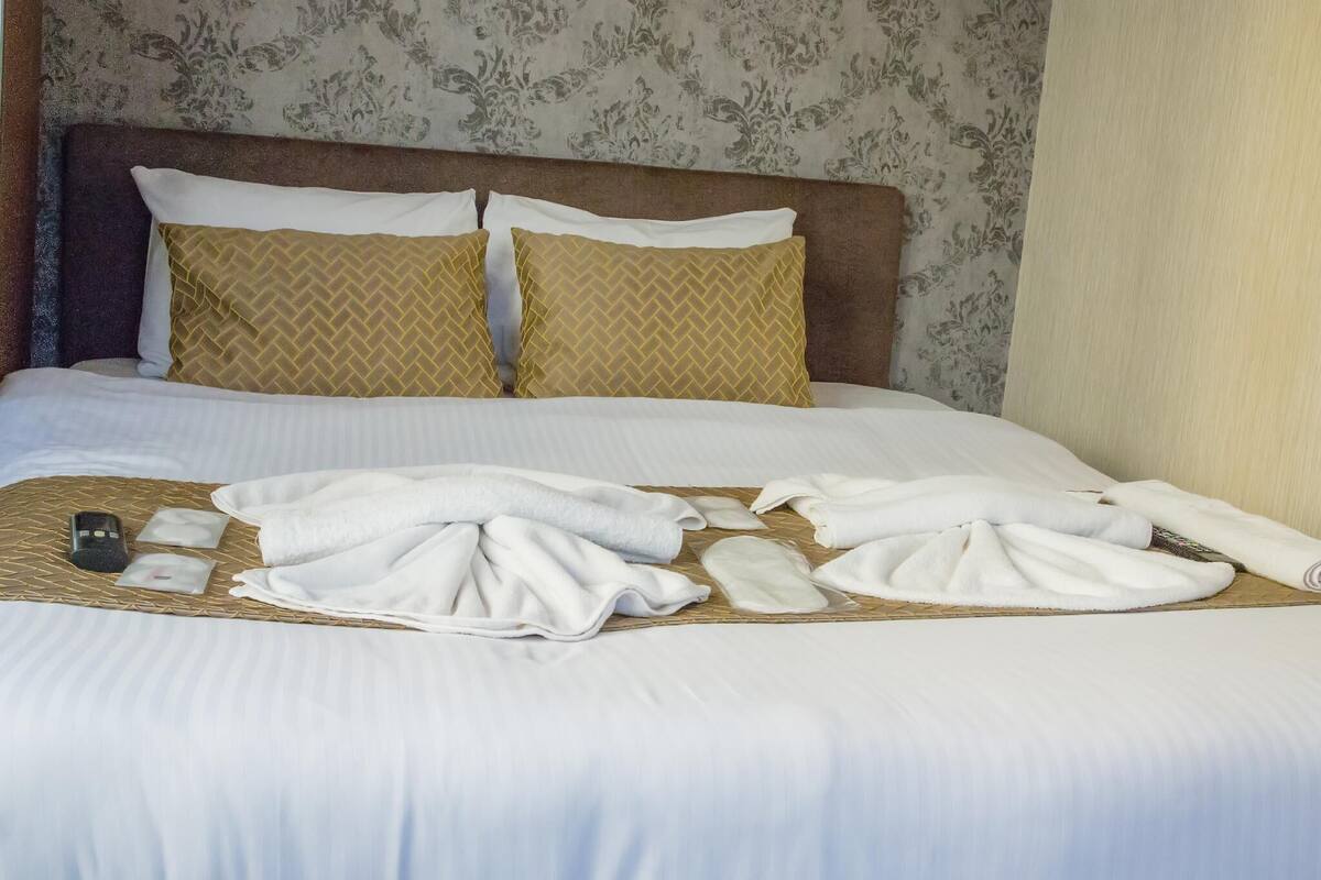 A neatly made bed with white linens, two gold-patterned pillows, and a remote control on the bed.
