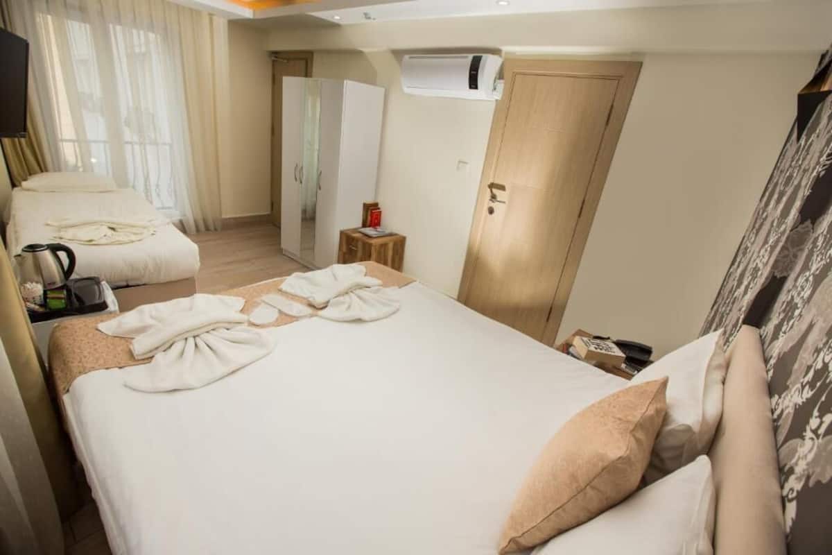 A hotel room with a bed, a chair, a wardrobe, an air conditioning unit, and a small table with a kettle and a book.