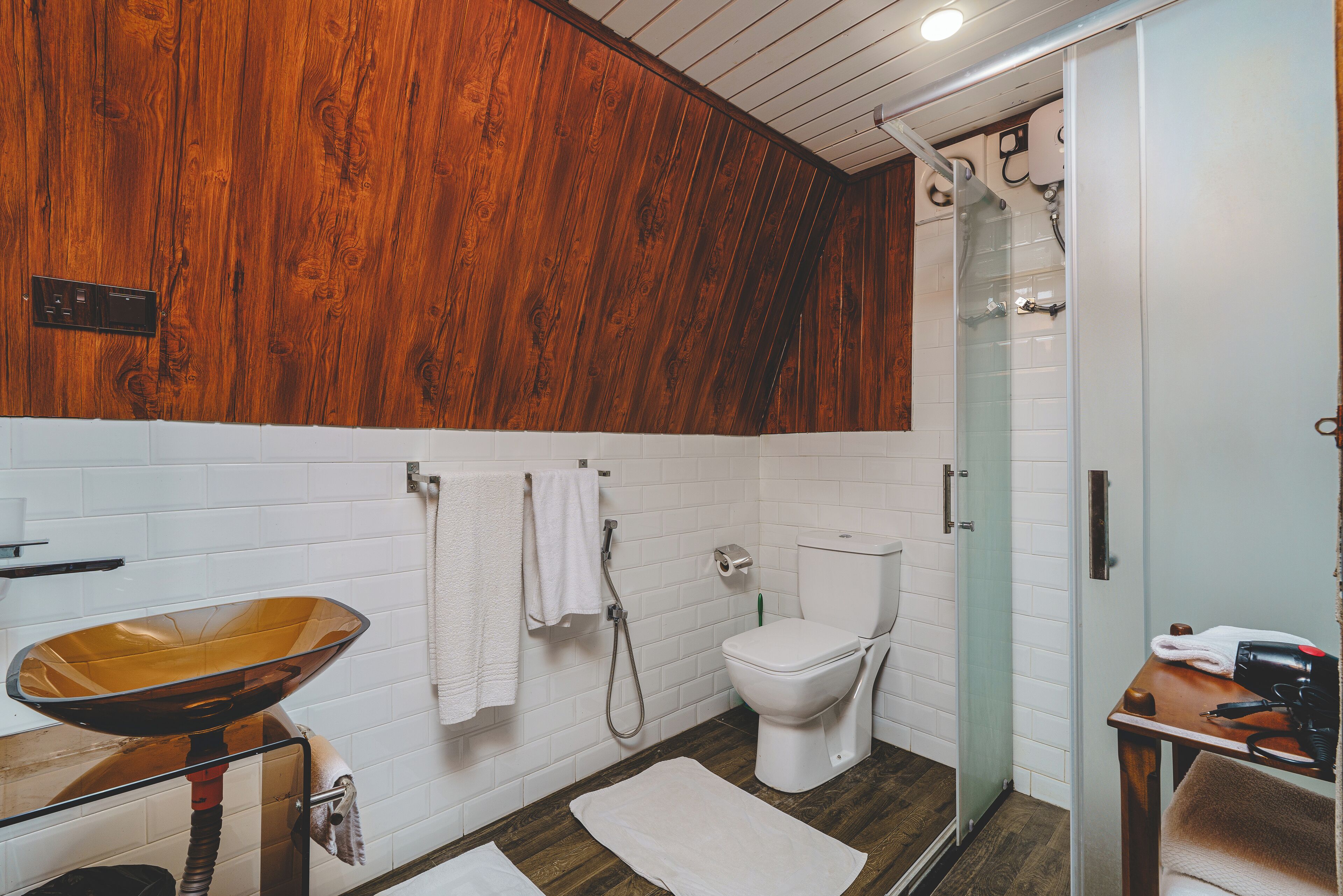 A bathroom with a wooden ceiling, white tiled walls, a wooden vanity with a bowl sink, a toilet, and a glass shower enclosure.