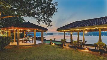 Two outdoor dining areas with tables and chairs, surrounded by potted plants and string lights, overlooking a calm body of water.