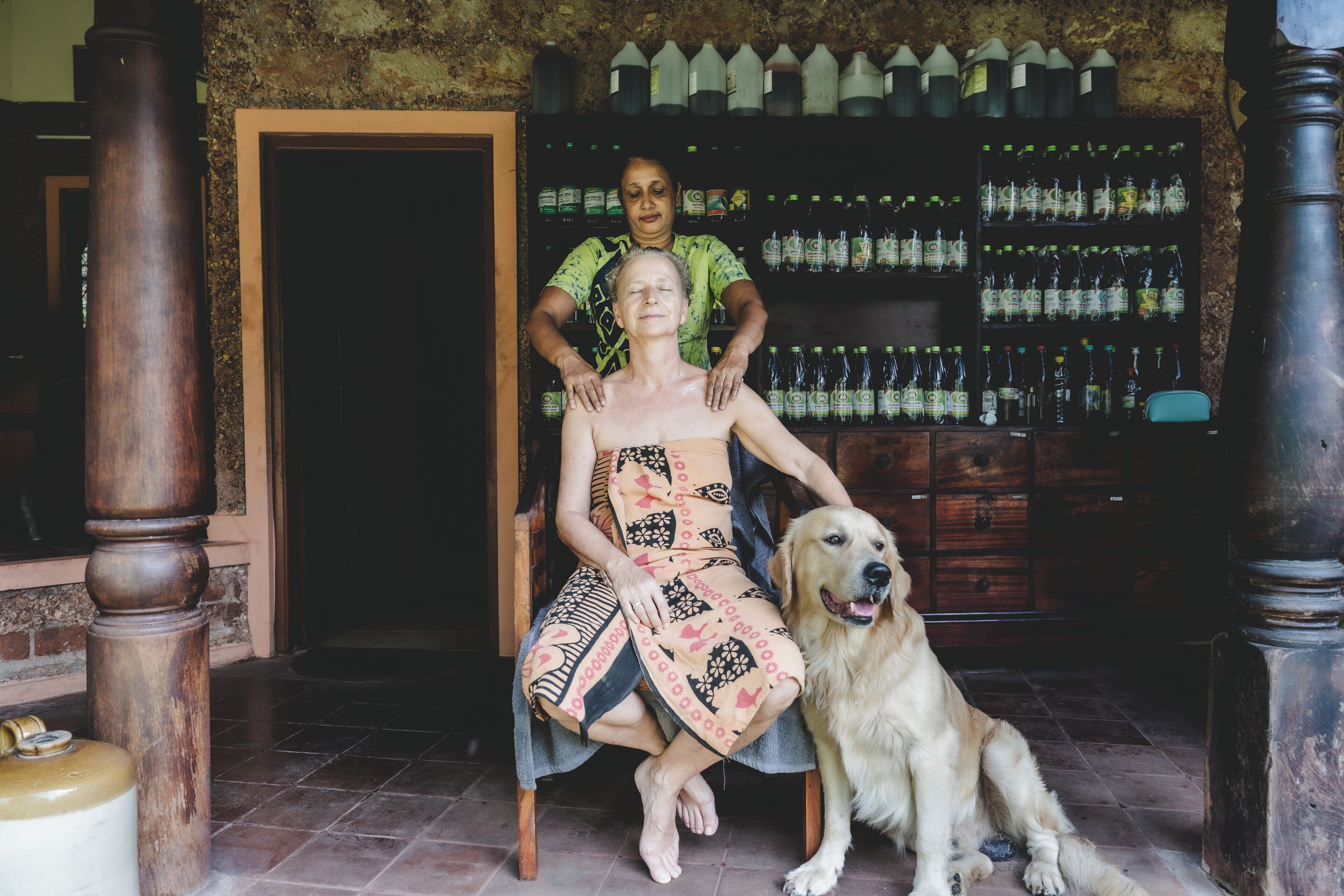 Una mujer sentada en una silla con un perro a su lado, en una habitación con piso de madera y una pared llena de bebidas embotelladas.
