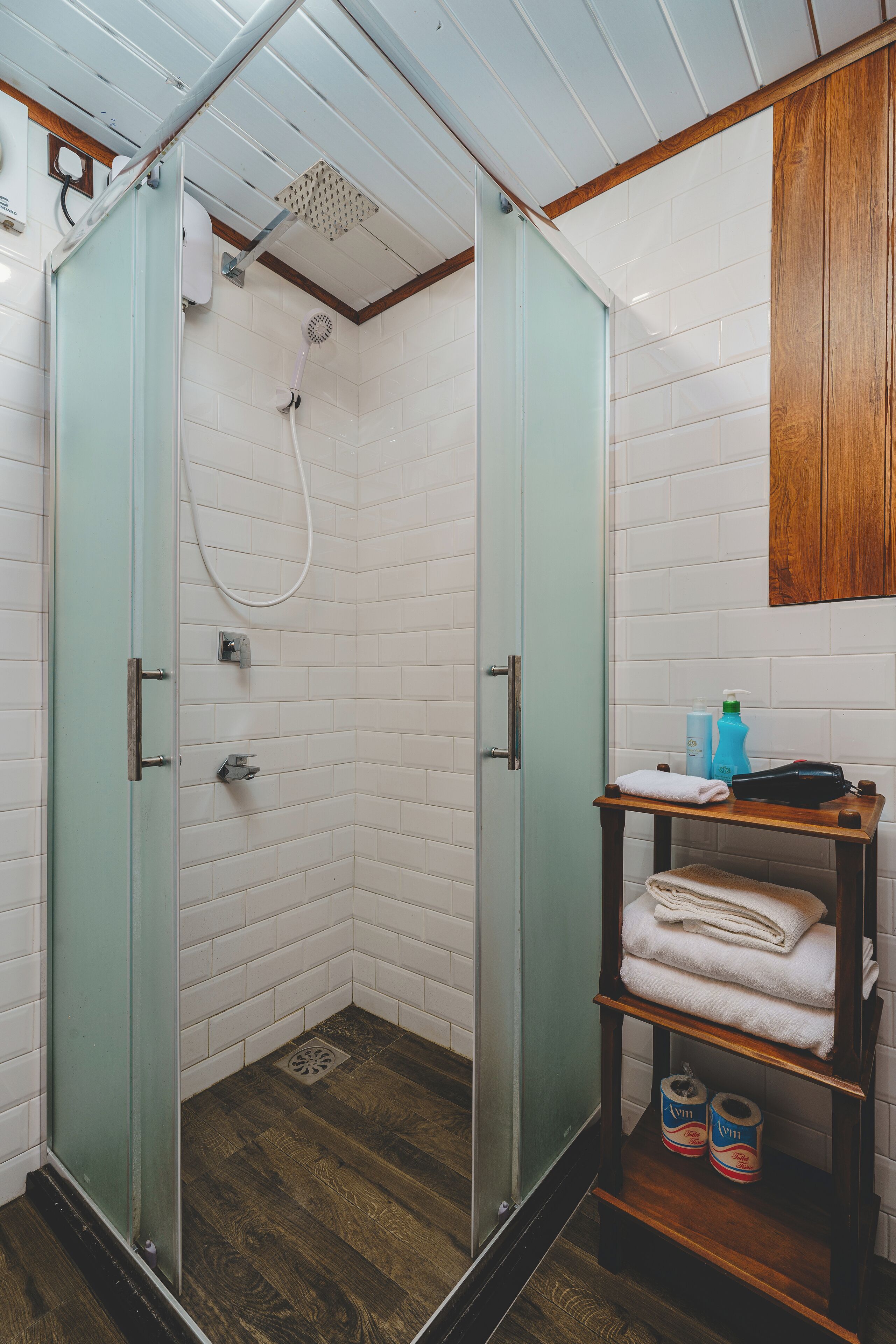 A bathroom with a glass-enclosed shower, white tiled walls, a wooden shelf with towels, and a wooden floor.
