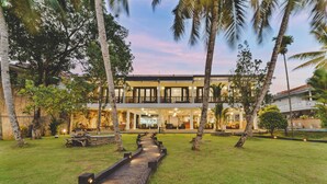 A two-story building with a central courtyard, surrounded by palm trees and a wooden pathway. - River View Villa (Dharga Town)