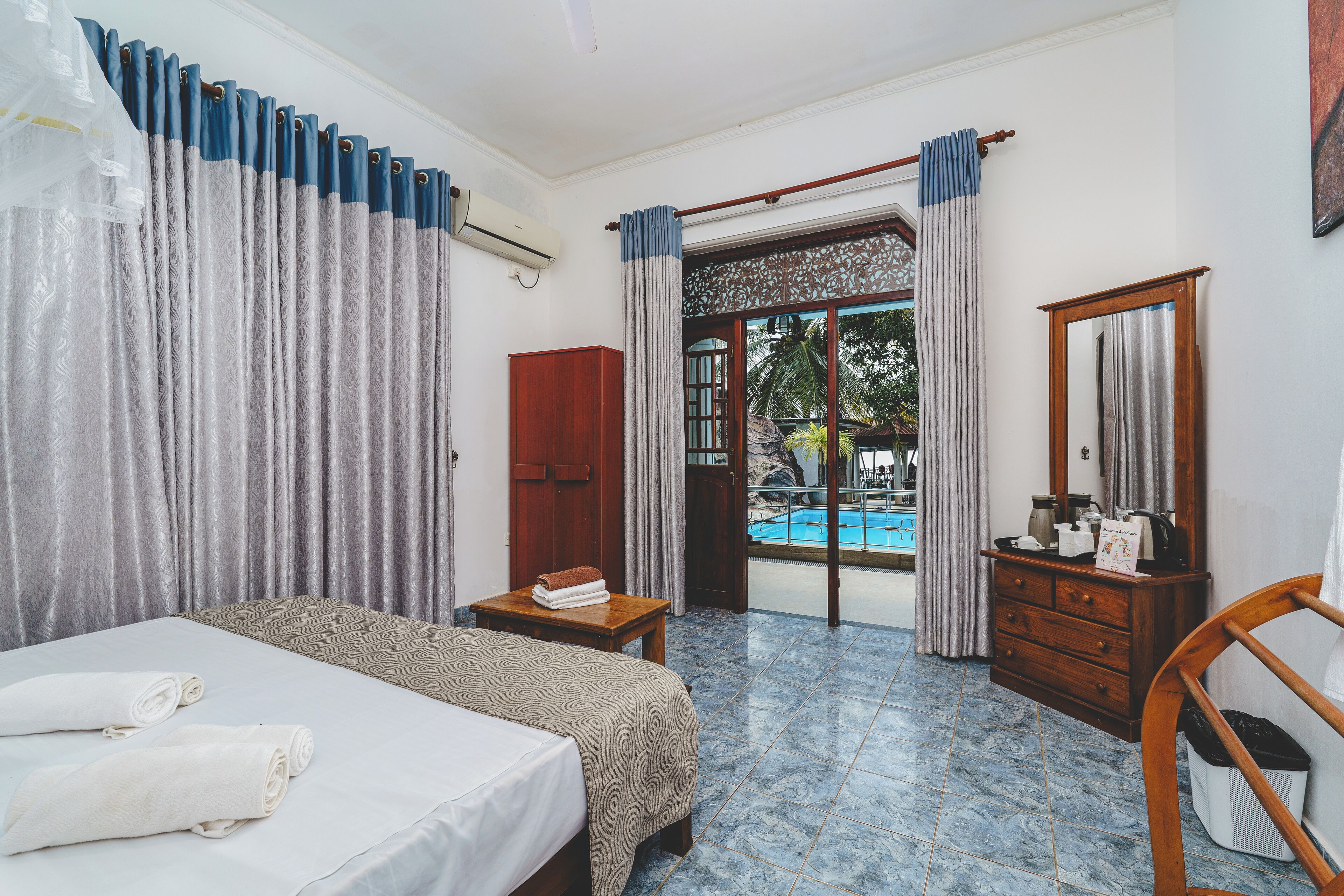 A hotel room with a bed, a wooden dresser, a small table with towels, a mirror, and a view of a swimming pool.