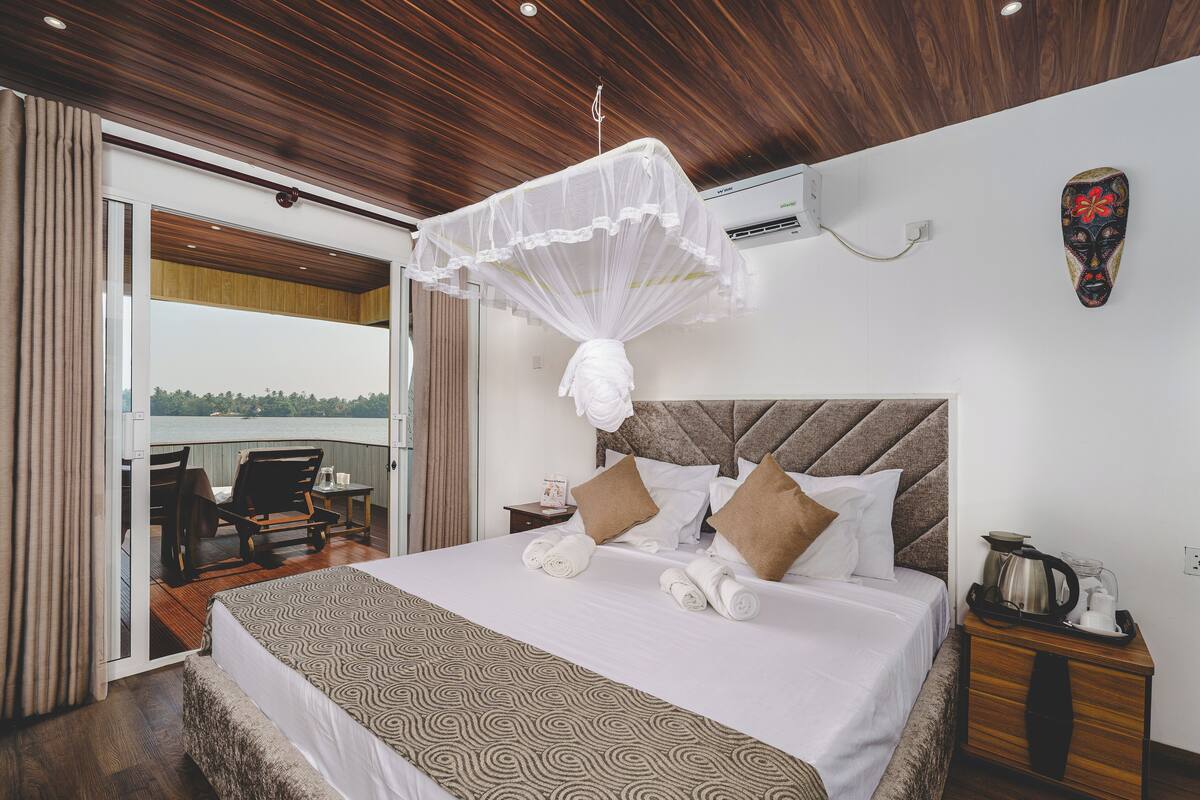A bedroom with a bed, a bedside table with a kettle, a view through a glass door, and a wooden ceiling.