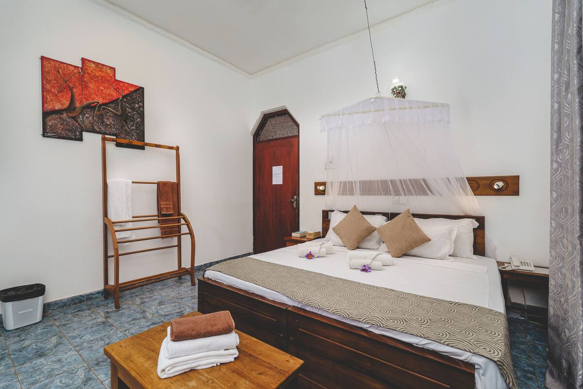 A hotel room with a bed, a wooden bench, a drying rack, and artwork on the wall.