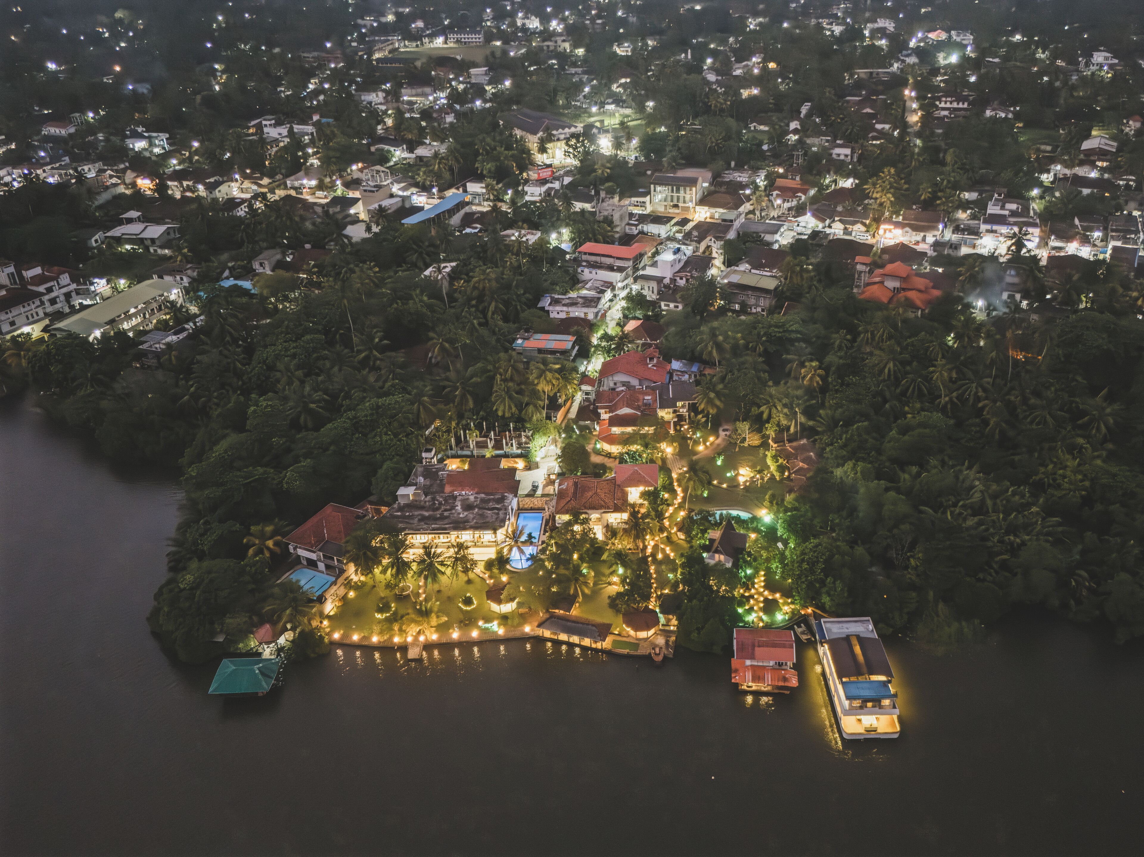An aerial view of a coastal town at night with illuminated buildings, a dock with boats, and a waterfront promenade.