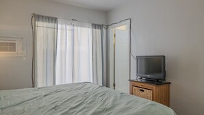 A bedroom with a bed, a television on a wooden cabinet, and a window with curtains. - Pool View King Suite #110 (New Smyrna Beach)