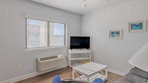 A living room with a TV on a white stand, a small table, a couch, and a chair. - Pool View King Suite #110 (New Smyrna Beach)