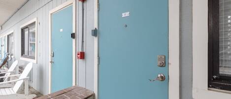 Una hilera de puertas azules con detector de incendios y cerradura electrónica, un banco de ladrillo y un ventanal con diseño de cuadrícula.