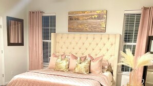 Un dormitorio con cabecera capitonada, cama con ropa de cama rosa, banco de madera, alfombra mullida y ventilador de techo.
