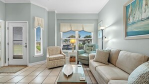 Un salon avec un canapé beige, une table basse en verre, un fauteuil à motifs et une vue sur l’extérieur grâce à de grandes fenêtres.