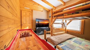 A cozy cabin interior with a wooden floor, a shuffleboard table, a bed with a quilt, a television, and a bunk bed with a view of the outdoors. - *Charming 4BD Lodge w/ Hot Tub! Near Town + GAMES* (Pigeon Forge)