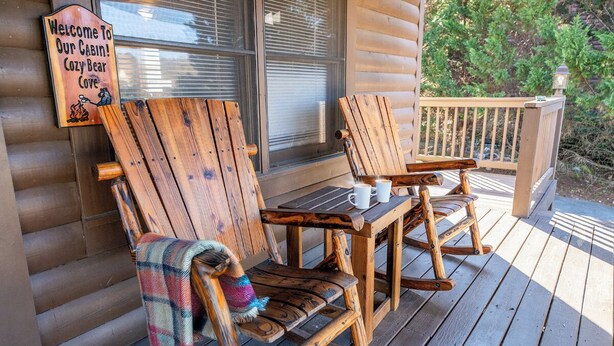 A wooden deck with two Adirondack chairs and a small table, a sign reading "Welcome to Our Cabin! Cozy Bear Cove", and a window with blinds. - *Charming 4BD Lodge w/ Hot Tub! Near Town + GAMES* (Pigeon Forge)