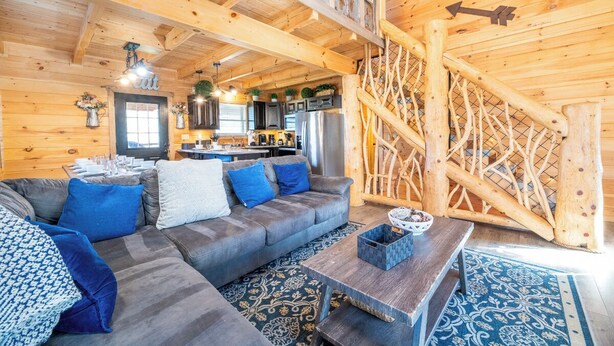 Un interior acogedor de cabaña con detalles de madera, un sofá con cojines azules, una mesa de comedor y una escalera con barandilla de madera.