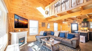 Interior acogedor de una cabaña de madera con chimenea, un sofá grande, un comedor y una cocina.