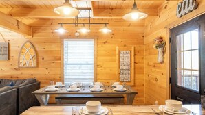 Interior de una cabaña de madera con una mesa de comedor preparada para cuatro comensales, un sofá y una ventana con persianas.