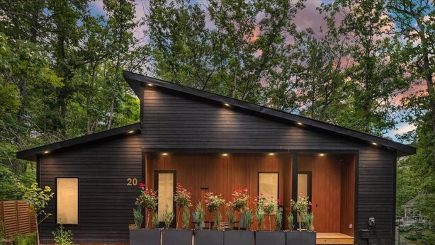 Une cabane moderne en bois, dotée d’un porche couvert, portant le numéro 20, entourée d’une allée en gravier et agrémentée de diverses plantes.