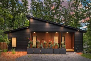 A modern wooden cabin with a covered porch, numbered 20, surrounded by a gravel driveway and landscaped with various plants.