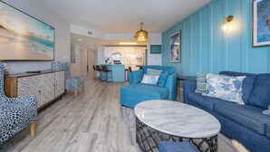 Un salon moderne avec un canapé, un fauteuil et une table basse, une cuisine en arrière-plan et un ventilateur de plafond.