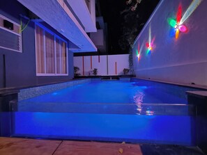 Une piscine éclairée de lumières bleues, entourée d’un bâtiment moderne doté de bandes lumineuses verticales et d’une fenêtre avec des rideaux.