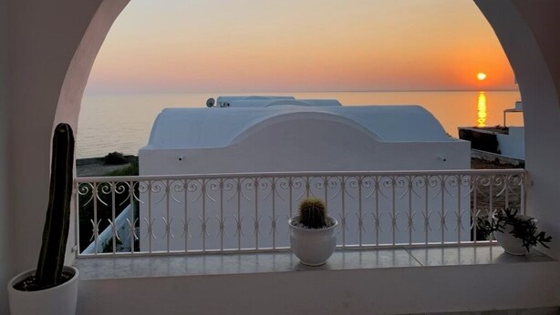 Een balkon met uitzicht op de ondergaande zon boven de zee, een witte reling en plantenbakken.