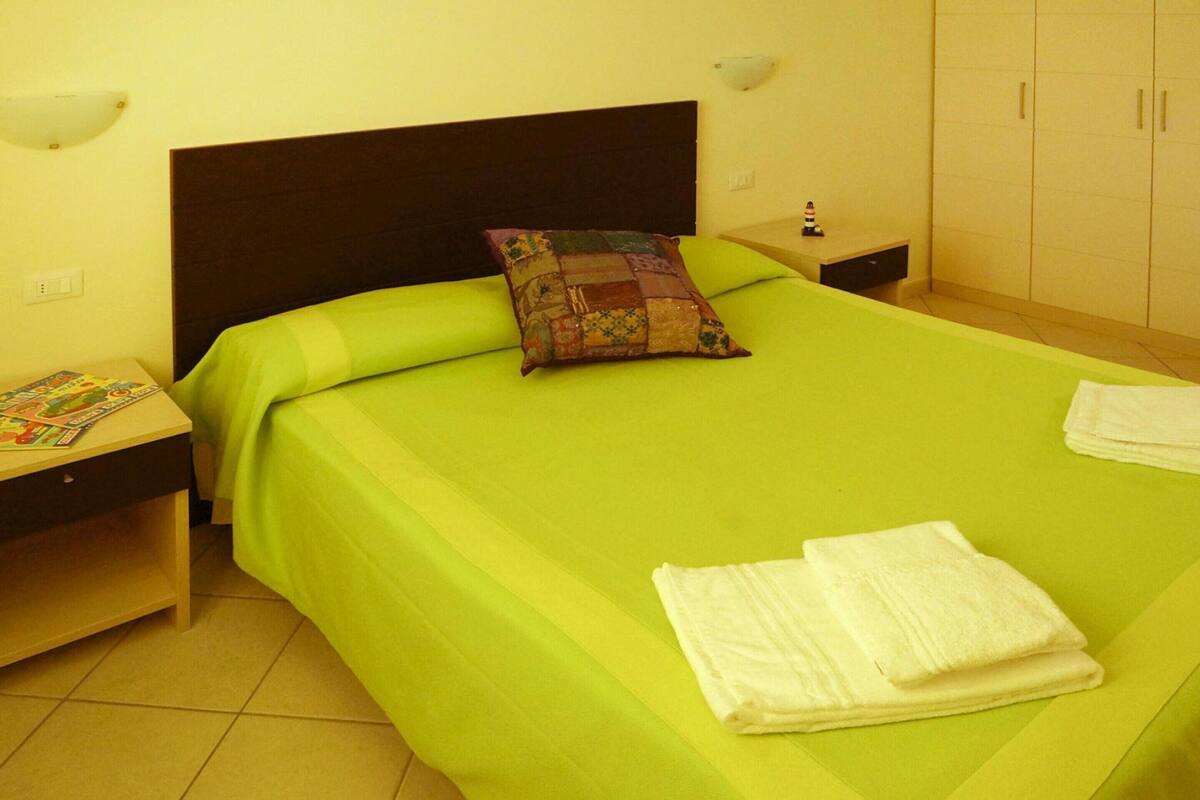 A hotel room with a green bedspread, a colorful pillow, and neatly folded white towels on the bed.