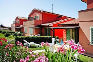 A multi-story residential building with a red facade, a covered patio area, and a well-maintained garden with pink flowers. - Apartment in Vada Near Restaurants (Rosignano Marittimo)