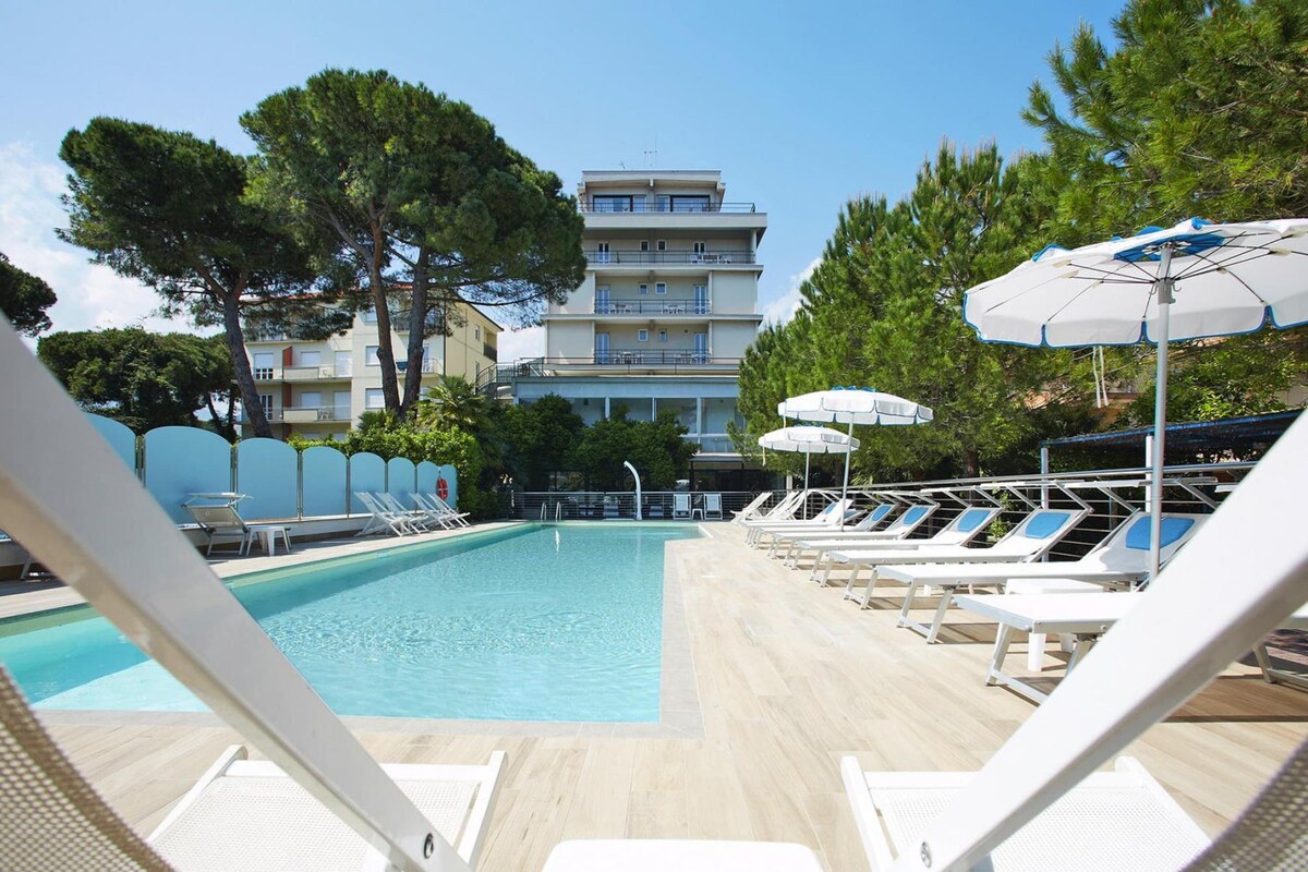 A swimming pool with lounge chairs and umbrellas, surrounded by trees and a multi-story building.