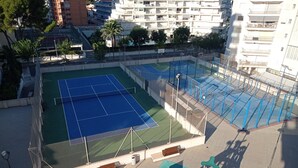 Ulkotenniskenttä, jonka ympärillä on aita, ja sen edessä on leikkialue.