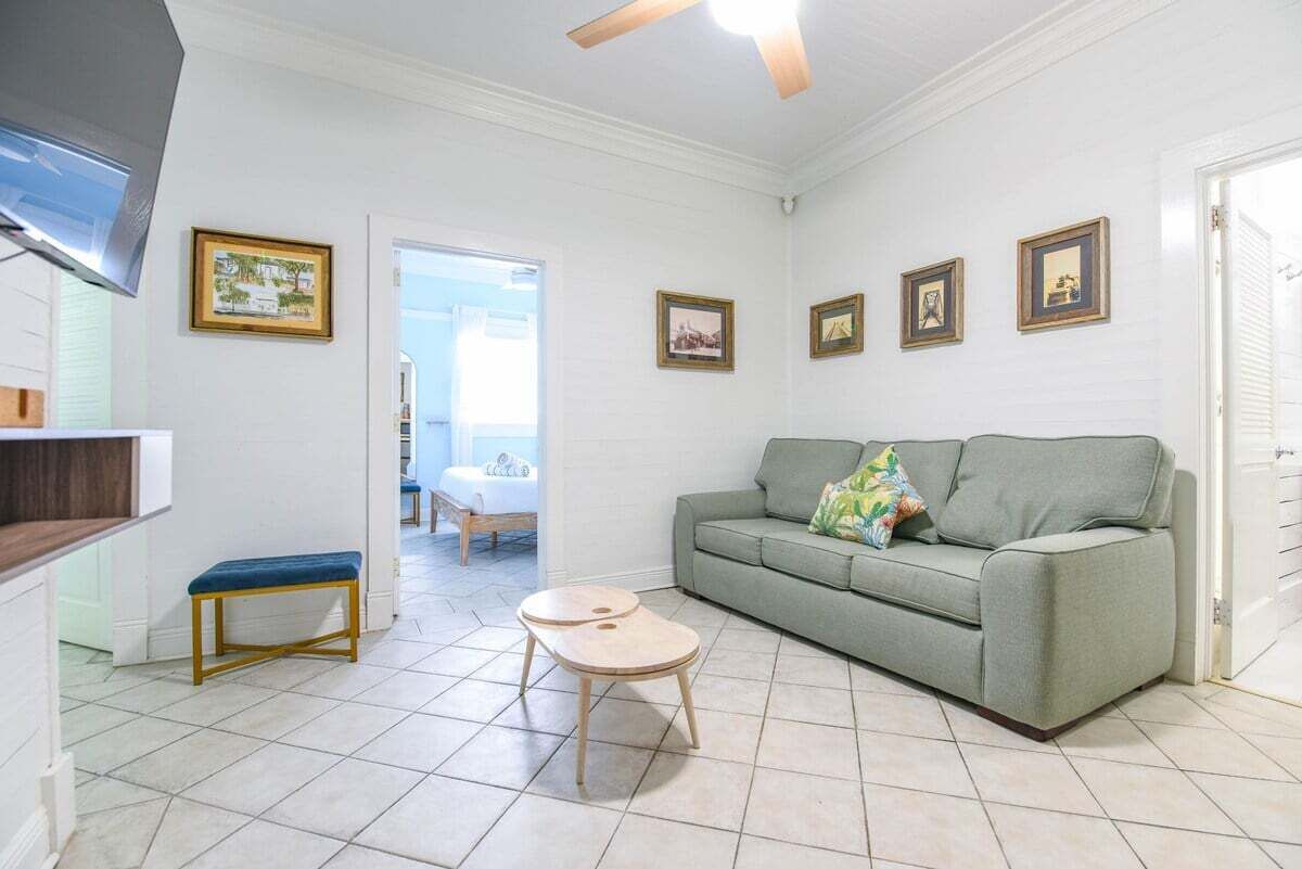 A living room with a green sofa, a wooden bench, and a small table. There are framed pictures on the wall and a ceiling fan.
