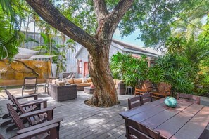 Un espacio de patio al aire libre con mobiliario de madera, un árbol grande y abundante vegetación.