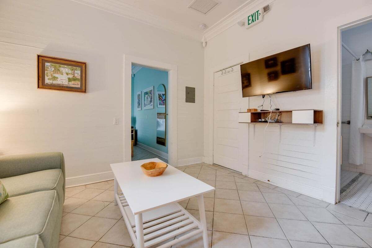Un hall de hotel con un sofá blanco, una mesita con un cuenco, un televisor montado en la pared y una puerta que da a otra habitación.