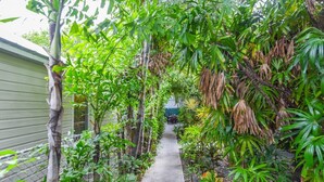 A narrow concrete pathway flanked by lush green plants and trees, leading to a building with a green exterior. - Atocha Cottage at Harbor Walk Hideaways | 2 Blocks From Duval! (Key West)