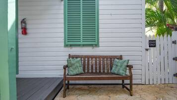 A wooden bench with patterned cushions in front of a white building with green shutters and a white picket fence.