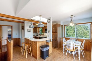A spacious kitchen with wooden cabinets, a central island with stools, pendant lighting, and a large window offering a view of greenery. - Classic Outer Banks soundside beach house near Duck Town Park! (Duck)