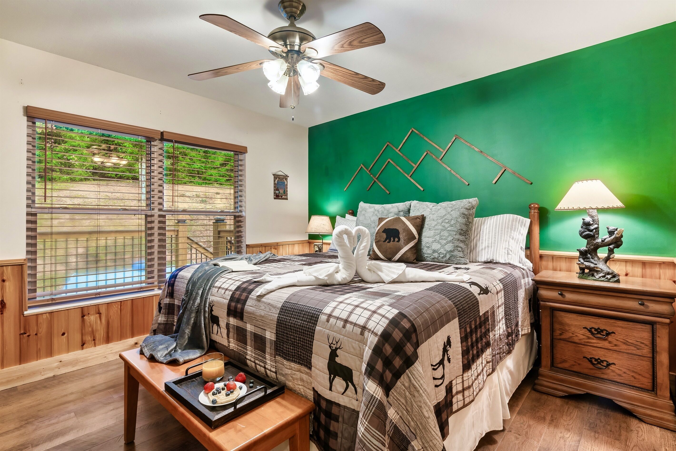 A bedroom with a green accent wall, a bed with a plaid quilt, a wooden nightstand, a ceiling fan, and a window with blinds.