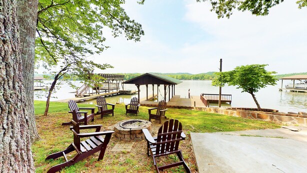 A lakeside outdoor seating area with Adirondack chairs and a fire pit, surrounded by a wooden deck and a gazebo.