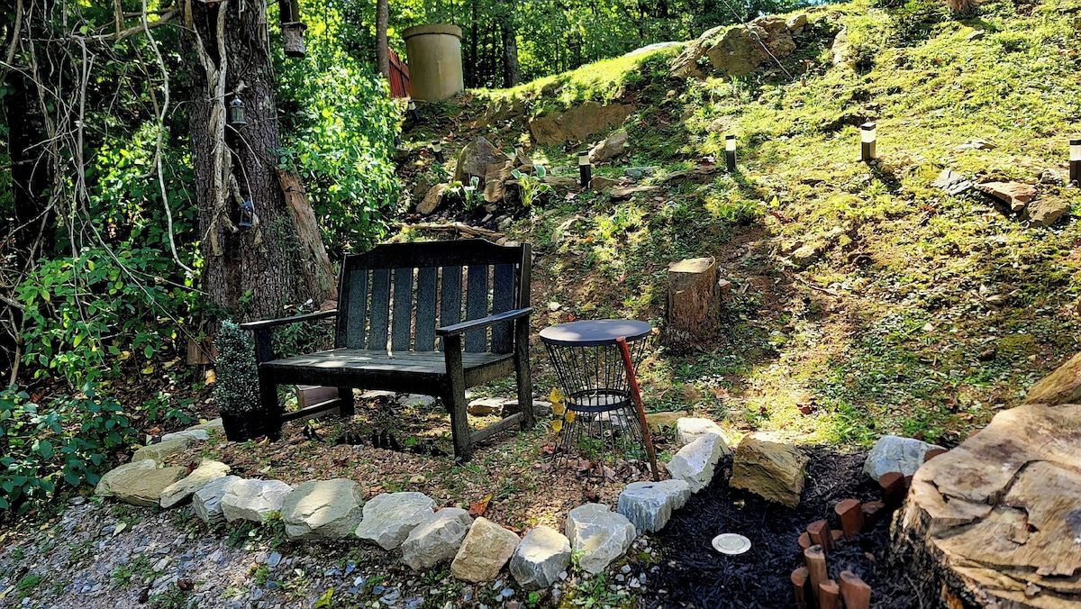 A bench and table in a natural setting with a stone border, surrounded by greenery and trees.