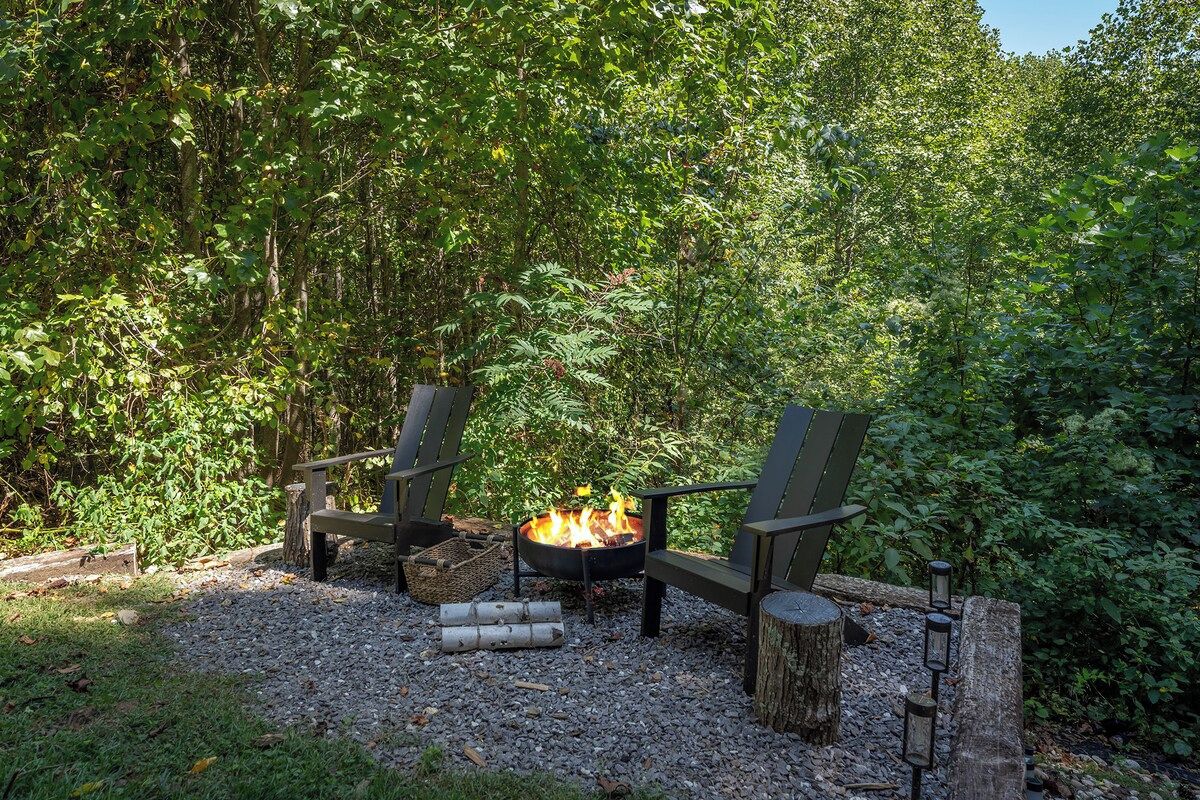 A fire pit with two Adirondack chairs and a log fire in a forested area.
